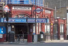 A red-bricked building with a rectangular, white sign reading "ENTRANCE" in black letters and a rectangular, blue sign reading "MAIDA VALE STATION"