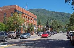 A wide undivided street with cars traveling in both directions going past a large brick building. In the rear is a forested mountain