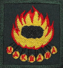 A green cloth badge with an embroidered camp fire. Each of the logs has one letter from the word Makwawa on its end