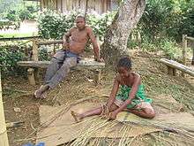 A Malagasy girls sits on the ground weaving with reeds, while an older gentleman sits on a bench above her.