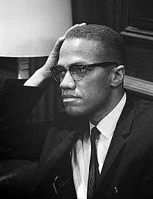 An African American man in his forties, wearing glasses and a suit and tie, sitting and looking to the right, with his hand resting on his right temple.