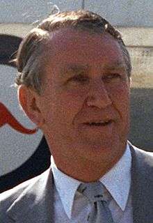 Photograph of a man aged about fifty, he has a weathered face and greying hair parted on the right. He wears a suit and tie; behind him can be seen part of a large aircraft with a kangaroo logo.