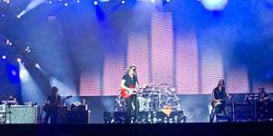 Five people. On the far left a person behind a console with a keyboard. Beside a person standing with semi-long hair, holding a bass guitar. Another person standing at the center, wearing black, holding a red guitar. To his right a person with long hair wearing black with a bass guitar in his hand. On the extreme right a person with a beret behind a drum kit.