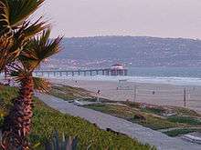 A photograph of a beach and a pier extending into the sea. In the foreground is a palm tree and a sidewalk, visible on the beach are a lifeguard station and a volleyball net, and on the horizon is a populated hillside.