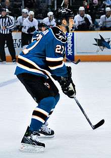 An ice hockey player, seen from the side, in a ready position. He is slightly crouched while standing on his skates and holds his stick in both hands. He wears a teal jersey with black trim, as well a black helmet.