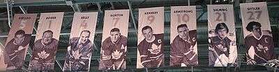 We are looking up at the ceiling of an arena where there are nine banners hanging. On each banner is the picture of a hockey player in a Toronto Maple Leaf uniform. Above each player's photo is a number and the player's name