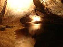 4-metre-wide cave passage, with a roughly triangular ceiling above head height, a wide, calm, brown stream running along the floor and a sandy bank on the left.