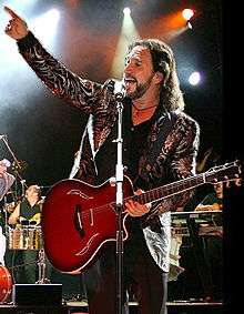 A man with a sparkly jacket on, with a guitar on his shoulder, and his right hand in the air
