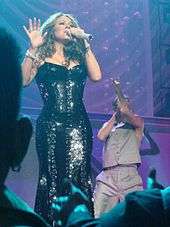A bold woman is singing on stage. She wears a black dress that has many mirrors. Behind her, a man with a brown suit is seen.