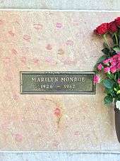 Photo of Monroe's crypt, taken in 2015. "Marilyn Monroe, 1926–1962" is written on a plaque. The crypt is covered in lipstick prints left by visitors and pink and red roses are placed in a vase attached to it.