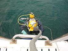 A diver wearing a lightweight helmet with surface supply umbilical and a single back mounted bailout cylinder is shown from above, partly in the water, climbing a boarding ladder on the side of a boat.