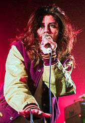 A young brunette woman wearing a baseball-style jacket, singing into a microphone against a pinkish background.