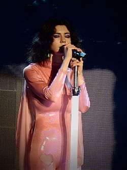 A young brunette woman in a long-sleeved pink dress, singing into a microphone