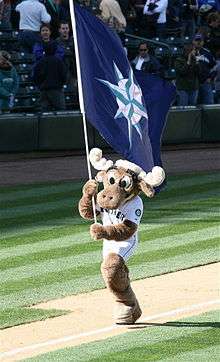 A large moose mascot runs down the third-base line of a baseball diamond. He is carrying a large navy blue flag adorned with a teal and silver compass.