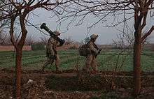  Two marines walking along the edge of a field, one of them carrying a Javelin missile tube