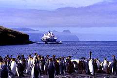 View of king penguins on a beach (foreground), a ship (middleground) and a distant, rugged island (background)
