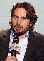 A brunette man is talking while he holds a microphone. He wears a blazer and a blue shirt. He has long hair and a beard.