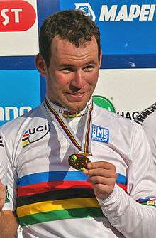 A man in his late twenties wearing a mostly white cycling jersey with rainbow stripes around the midsection and biceps. He has a medal hung around his neck, and clutches it with the fingertips of his left hand. He is smiling.
