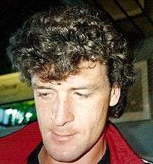 A head shot of a young man with curly long brown hair that is greying slightly. He is looking down, away from the camera and the red collar of a jacket is visible.