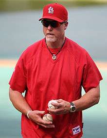 A man in a red athletic shirt holds a baseball in each hand. He is wearing a red cap and sunglasses.