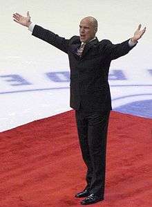 A bald man extends his hands to spectators at a hockey game. He is wearing black clothes and is standing on a red carpet.
