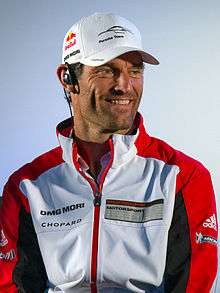 A man in his late thirties wearing a red, white and grey jacket and a white baseball cap with sponsors logos.