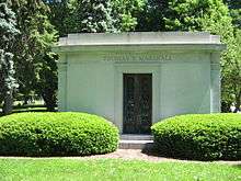 A large stone tomb with a metal gate in the center of the tomb, and with carved columns at the corners meeting a crest which circles the top of the building. Two large evergreen bushes obscure the lower portion of the tomb which sits in a grassy meadow.