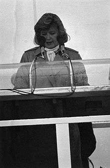A black-and-white photo of a woman in her fifties making a speech behind a podium and microphone