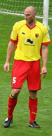 A man wearing a yellow shirt, red shorts, red socks and black football boots, standing on a grass field