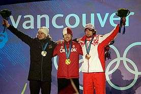 Three young men dressed in winter clothing stand embraced side-by-side in front of a wall with the Olympic rings and the word Vancouver. The man on the center carries a gold medal around his neck, while the two men on the left and on the right carry a silver and a bronze medal, respectively, and hold up a flower bouquet.