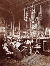 A black and white photograph of an elderly Victoria sat alongside a younger woman (Beatrice) reading a newspaper. The room is ornately decorated, with a number of photographs, paintings and a large chandelier hanging from the ceiling.