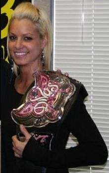 A blonde Caucasian woman holding a wrestling championship on her left shoulder. She is wearing a black, long-sleeve T-shirt and long silver earrings. The championship belt has a black strap, and the front of the belt is in a pink butterfly design, with the word "Divas" in white.