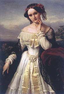 A three-quarter length portrait of a young white woman in the open air. She wears a shawl over an elaborate long-sleeved dress that exposes her shoulders and has a hat on over her centrally parted dark hair.