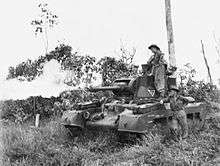 Black and white photo of a tank with flames coming out of the gun barrel on its turret. Two men wearing military uniforms are leaning on the side of the tank.