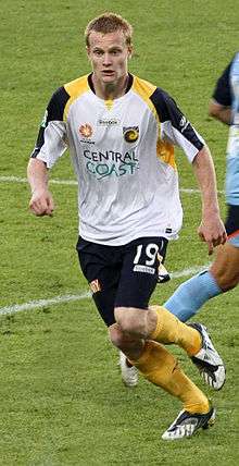 A man with light-coloured hair wearing a white shirt and navy shorts running forwards on a football field.