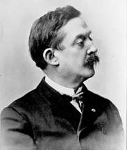 A man with a bushy mustache looks to the right while wearing a dark suit