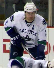 An ice hockey player leans forward with his stick. He has a serious look of concentration on his face as he looks into the distance. He wears skates, gauntlets, a helmet with a visor and a sweater with a stylized "C" shaped like a whale. Above the logo is the word "Vancouver," and a large "A" directly under his left shoulder.