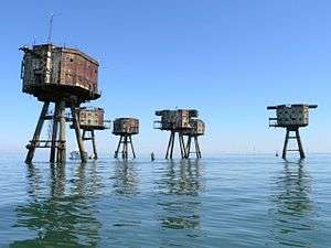 The view from a boat of a site containing six sea forts. The forts have an octagonal shape, with rusty metal walls and two rows of windows. Each fort is supported by four legs jutting from the sea at an angle.