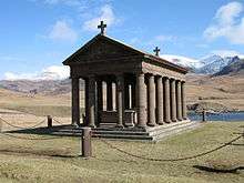 A stone building, with open sides lined by columns sits in a grassy meadow. A low fence made of small posts and a single chain surrounds it. Tall snow-clad mountains in the background lie under a blue sky.