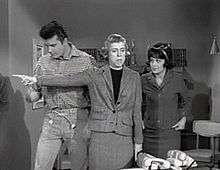 A black and white screenshot from the television series, The Beverly Hillbillies shows Max Baer, Jr. as Jethro, Nancy Kulp as Jane Hathaway, and Sharon Tate as Janet Trego, a secretary. Tate is wearing a business suit and a dark wig, and is watching Miss Hathaway