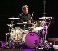 Gray haired man in a black shirt sitting behind a drumset