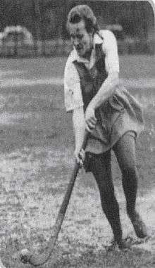 black-and-white photo of a female hockey player wearing a knee-length skirt and elbow length shirt. She is wearing dark colour tights. She is standing upright while holding the hockey stick pointed down at the ground.  She is in a running position and about to hit a ball that is shown in the lower left hand corner.