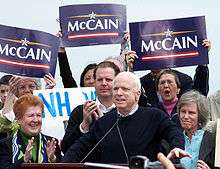 White-haired man speaking at podium, with group of people behind him, some holding blue "McCain" signs