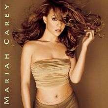 Image shows Carey standing in front of a brown/gold background in a beige sleeveless and mid-baring top, with darker matching pants. Her hair is long and golden-auburn, and is flowing in the air. her left hand is touching the flowing tips of her hair. She has a jeweled belt along her naval, with the words "Mariah Carey" written along the album cover.