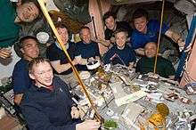 Nine astronauts seated around a table covered in open cans of food strapped down to the table. In the background a selection of equipment is visible, as well as the salmon-coloured walls of the Unity node.