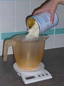 A cream-coloured powder is poured from a tin into a measuring jug on an electronic kitchen scale.