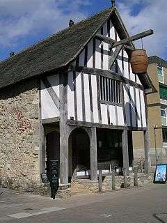 A photograph of a medieval building with white rendering and plain woodwork; a barrel is hung above the entrance and a small visitor's sign is placed on the street alongside the building.