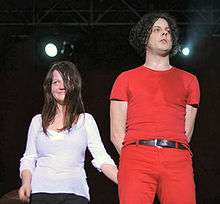 The White Stripes standing on stage: Meg is to the left, wearing a white shirt and black pants, smiling at the crowd; to her left is Jack, wearing a red outfit with a black belt.