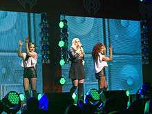 A young long-haired blonde woman singing into a microphone onstage. She sports a black skirt and black Bad Gal jacket. On her left and right are two brunette women dancing, each are wearing a sleeveless white top and leather shorts. A portraits of several bass speakers squared in pattern with background colors of neon green as the women's backdrop, as well as the iHeart Radio logo.