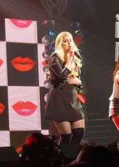A young long-haired blonde woman singing into a microphone onstage. She wears a black skirt and jacket. Images of several red lips appear behind the woman.